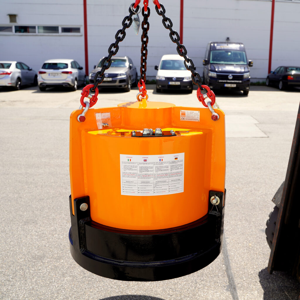 Ein orangefarbener Hydraulikmagnet hängt an schwarzen Ketten vor einer Werkhalle, während im Hintergrund mehrere geparkte Fahrzeuge unscharf zu sehen sind.