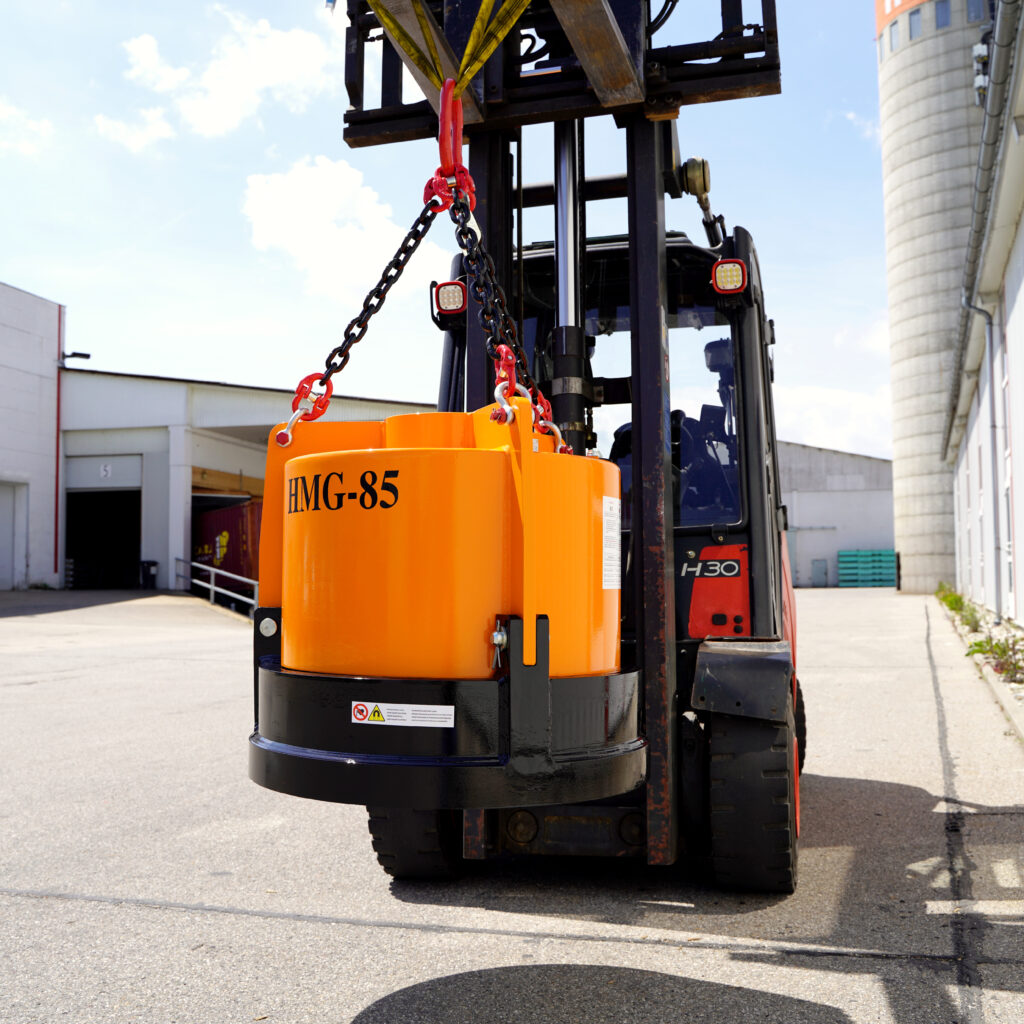 Ein orange-schwarzer Hydraulikmagnet HMG-85 hängt an Ketten vor einem Gabelstapler; im Hintergrund stehen Industriegebäude und blauer Himmel.