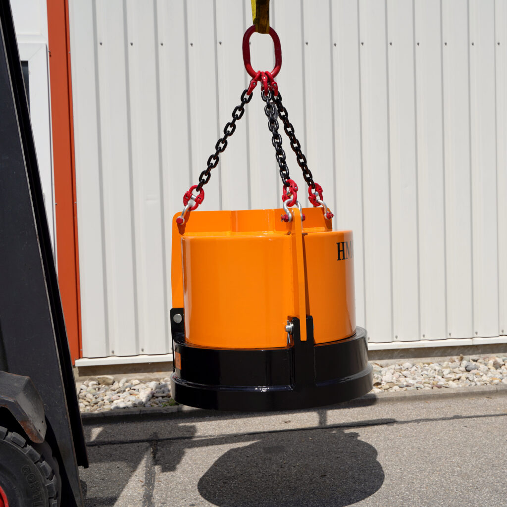 Ein orange-schwarzer Hydraulikmagnet hängt an Ketten vor einer hellen Metallwand, während links ein Teil eines Fahrzeugs ins Bild ragt.