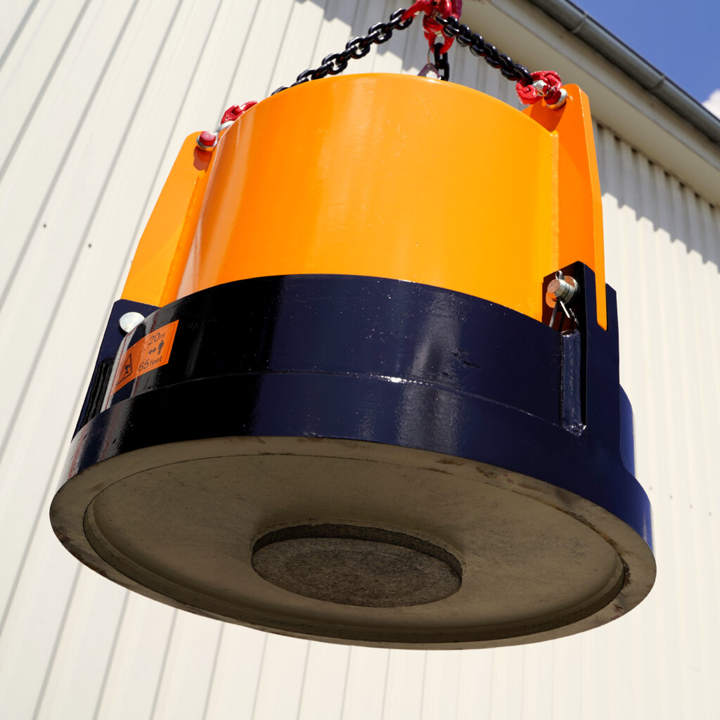 Ein orange-schwarzer Hydraulikmagnet hängt an Ketten vor einer hellen Metallfassade, darunter ist die runde Magnetfläche im Gegenlicht zu sehen.