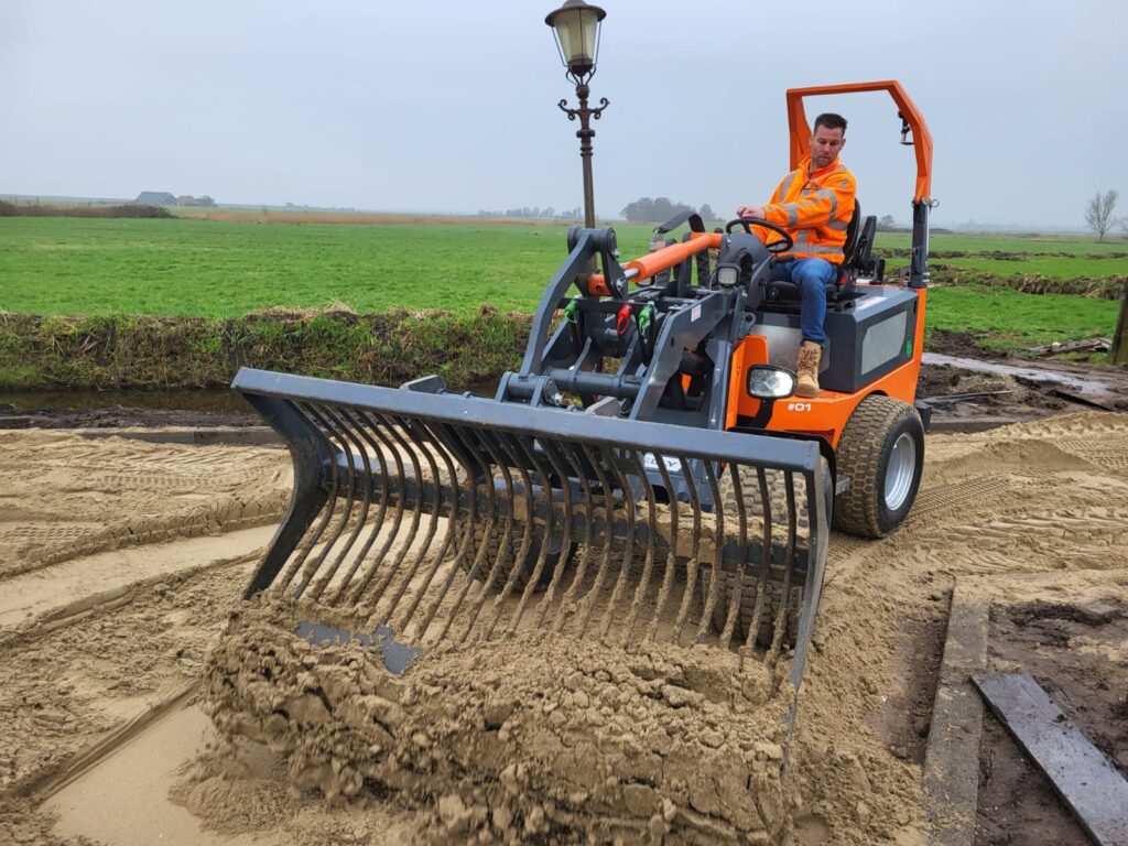 Ein Mann im orangefarbenen Warnanzug steuert eine orange-graue Baumaschine, die mit einer breiten Rechenvorrichtung Sand auf einem Feld verteilt.