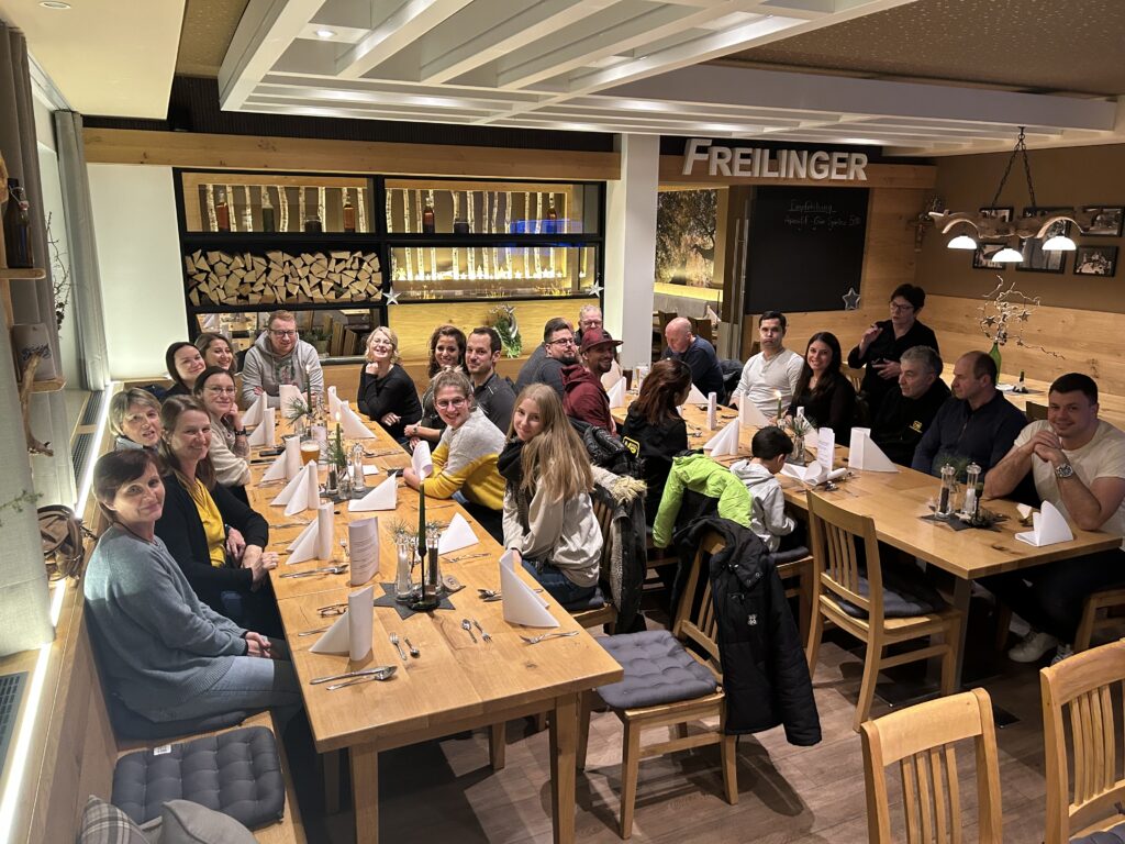 Eine große Gruppe sitzt in einem gemütlichen Restaurant mit Holzinterieur und dem Schriftzug Freilinger an der Wand, während sie gemeinsam am Tisch isst.
