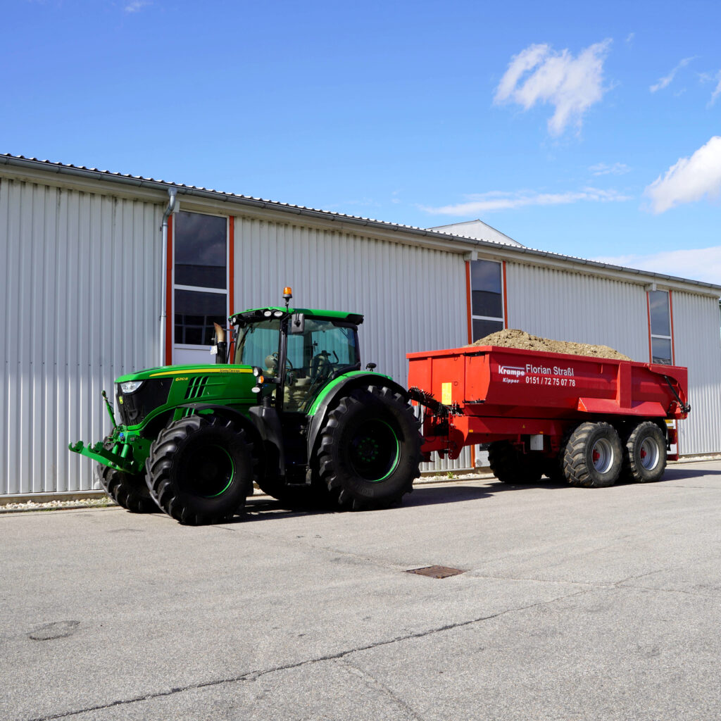 Ein grüner John Deere 6210R zieht einen roten Krampe-Kipper mit Erdhaufen vor einer hellen Industriehalle unter blauem Himmel über den Hof.