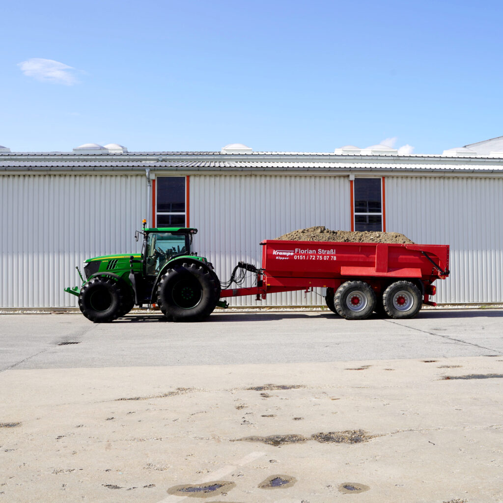 Ein grüner John Deere Traktor steht vor einer hellen Industriehalle und zieht einen roten Kippanhänger, der mit Erdmaterial gefüllt ist.