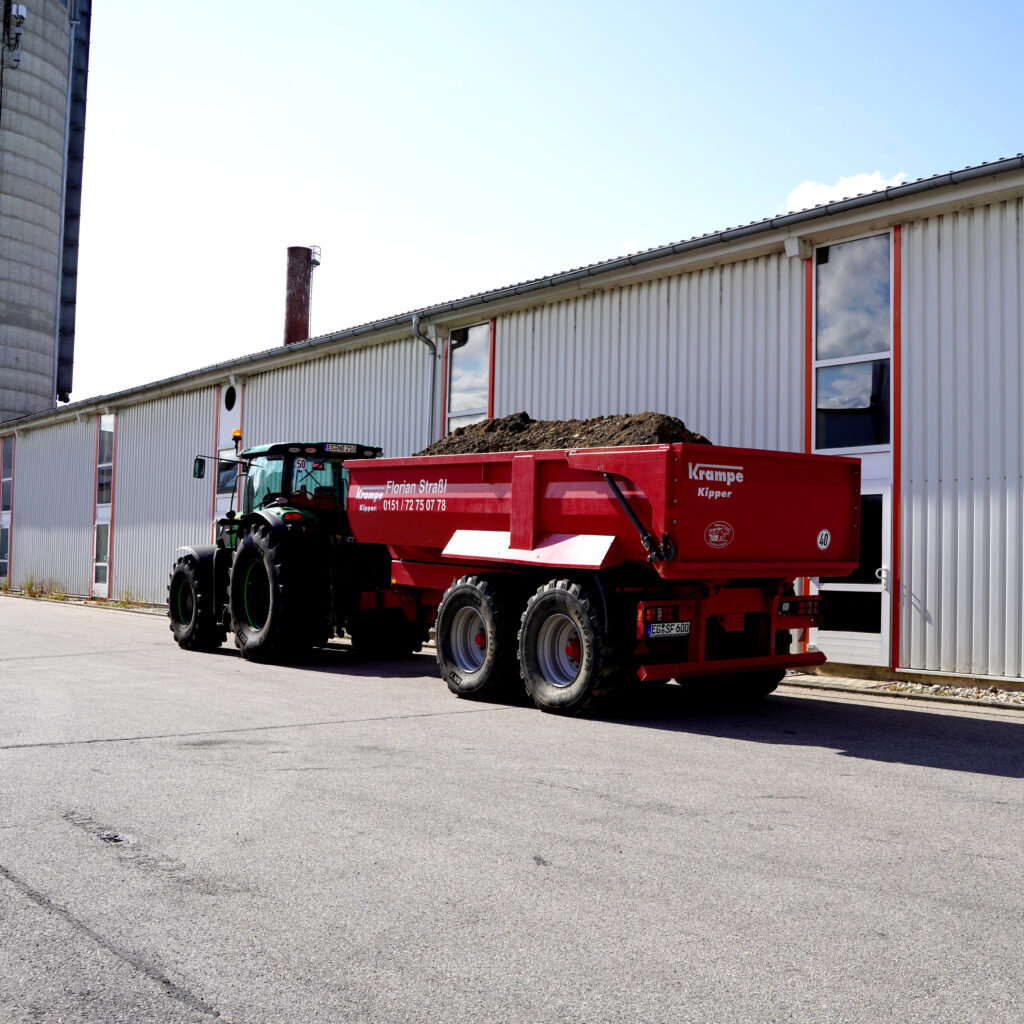 Ein grüner JohnDeere zieht einen rot beladenen Krampe-Kipper vor einer langen Industriehalle, darüber ein heller Himmel mit etwas Wolken.