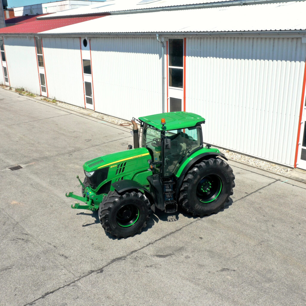 Ein grüner John Deere 6210R Traktor mit breiten Reifen steht vor einer hellen Industriehalle auf einem sonnigen Hof mit Asphaltboden.