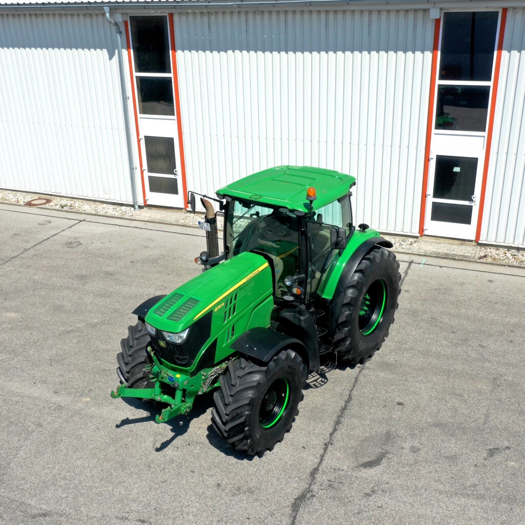 Ein grüner John Deere Traktor mit großen Reifen steht vor einer hellen Industriehalle mit roten Türrahmen auf dem Asphalt im Sonnenschein.