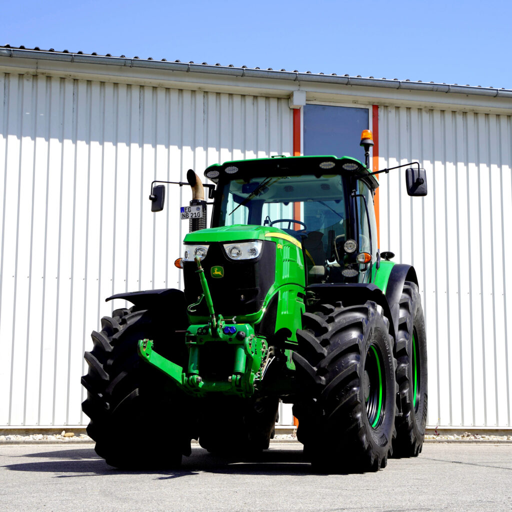 Ein grüner John-Deere-Traktor steht vor einer weißen Hallenwand auf dem Hof, mit großen Reifen, Warnleuchte und breiter Kabine im Sonnenschein.