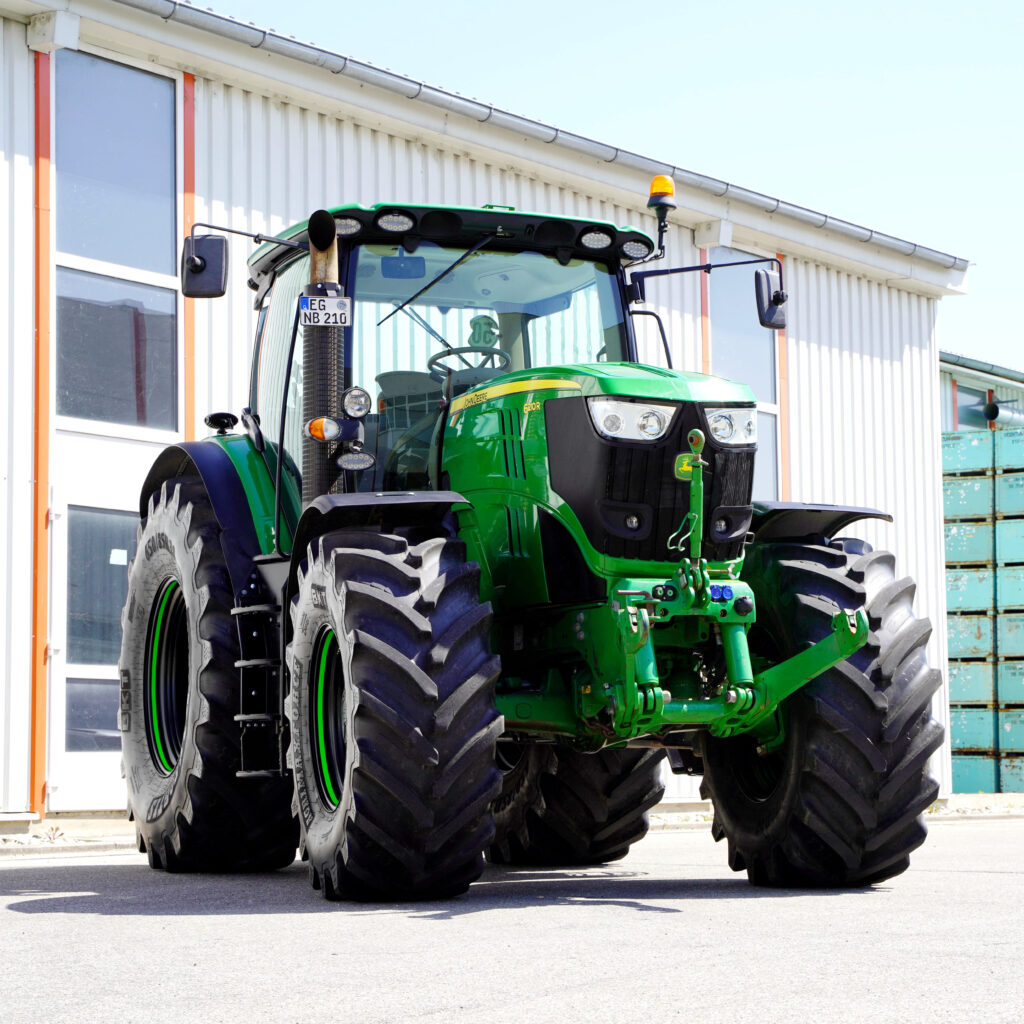 Ein grüner John Deere 6210R mit breiten Reifen, Frontanbau und gelber Rundumleuchte steht vor einem hellen Industriegebäude im Sonnenlicht.
