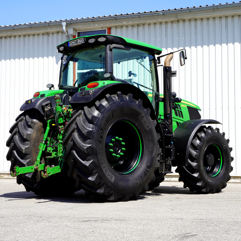Ein grüner John Deere 6210R steht vor einer weißen Hallenwand; große Bereifung, breite Kabine und schwarze Kotflügel prägen das Erscheinungsbild.
