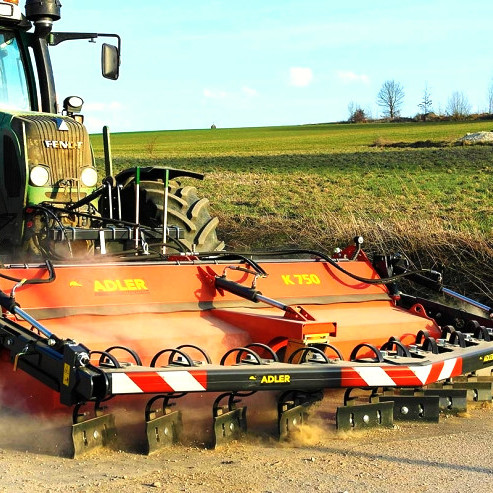 Auf einem Feldweg zieht ein Fendt-Traktor mit orangefarbener ADLER K750 Kehrmaschine Staub und Schmutz auf, während im Hintergrund grüne Felder liegen.