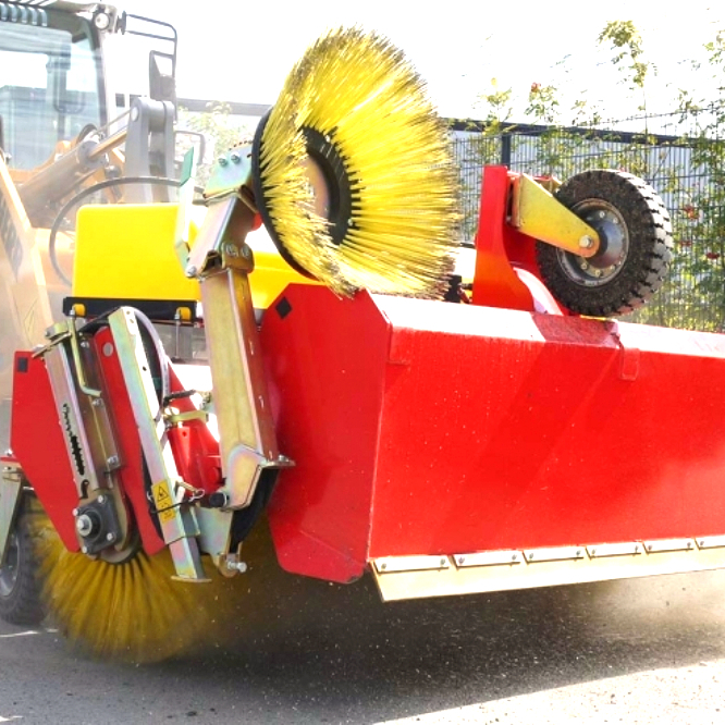 Eine rot-gelbe Kehrmaschine mit großen Bürsten und Stützrad arbeitet auf Asphalt vor einem Zaun, während feiner Staub in der Sonne aufwirbelt.