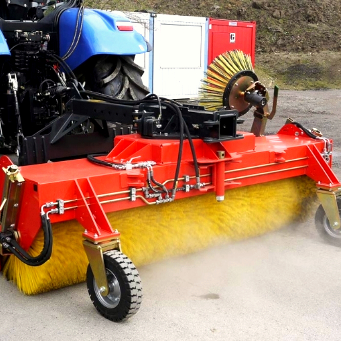 Ein roter Kehrbesen mit gelber Walzenbürste arbeitet an einem blauen Traktor, während feiner Staub über den Asphalt aufwirbelt.