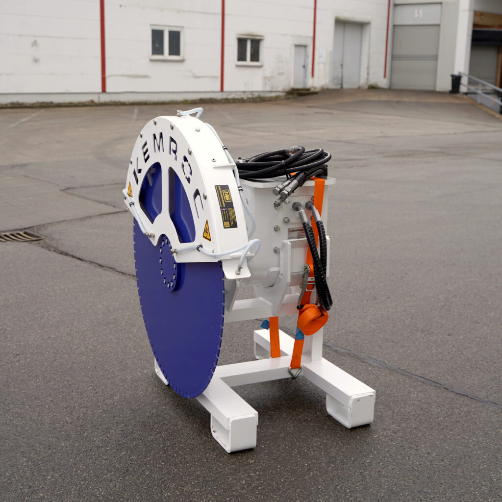 Vor einer Industriehalle steht ein blau-weißer Kemroc-Sägeschnittkopf mit angeschlossenen Hydraulikschläuchen und orangefarbenem Spanngurt.