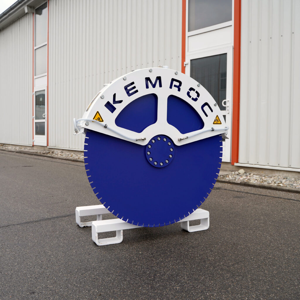 Ein blau-weißes Kemroc-Steinsegenrad steht auf dem Hof vor einer hellen Industriehalle mit orangefarbenen Rahmen und Warnsymbolen.