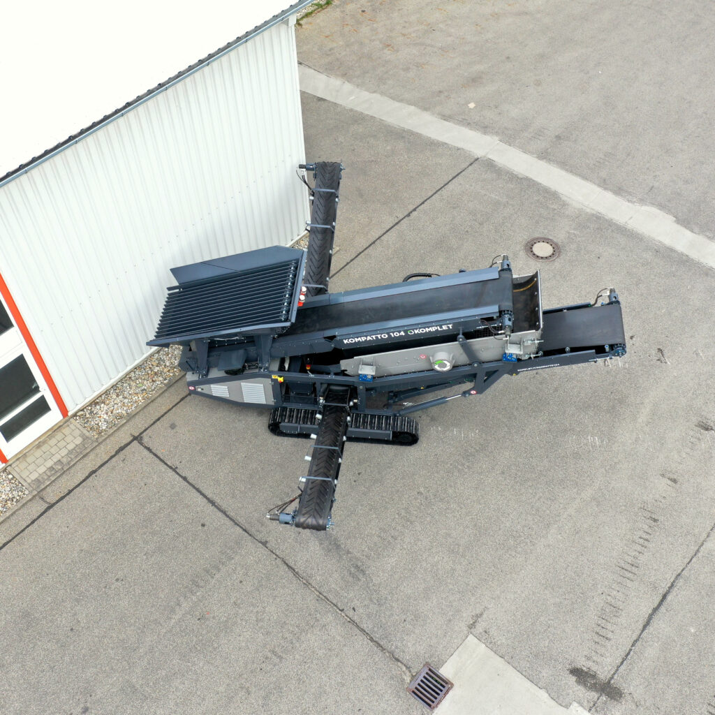 Ein schwarzer Kompaktor Kompatto 104 mit langen Förderbändern steht auf einem Asphaltplatz neben einer weißen Hallenwand und ist von oben aufgenommen.