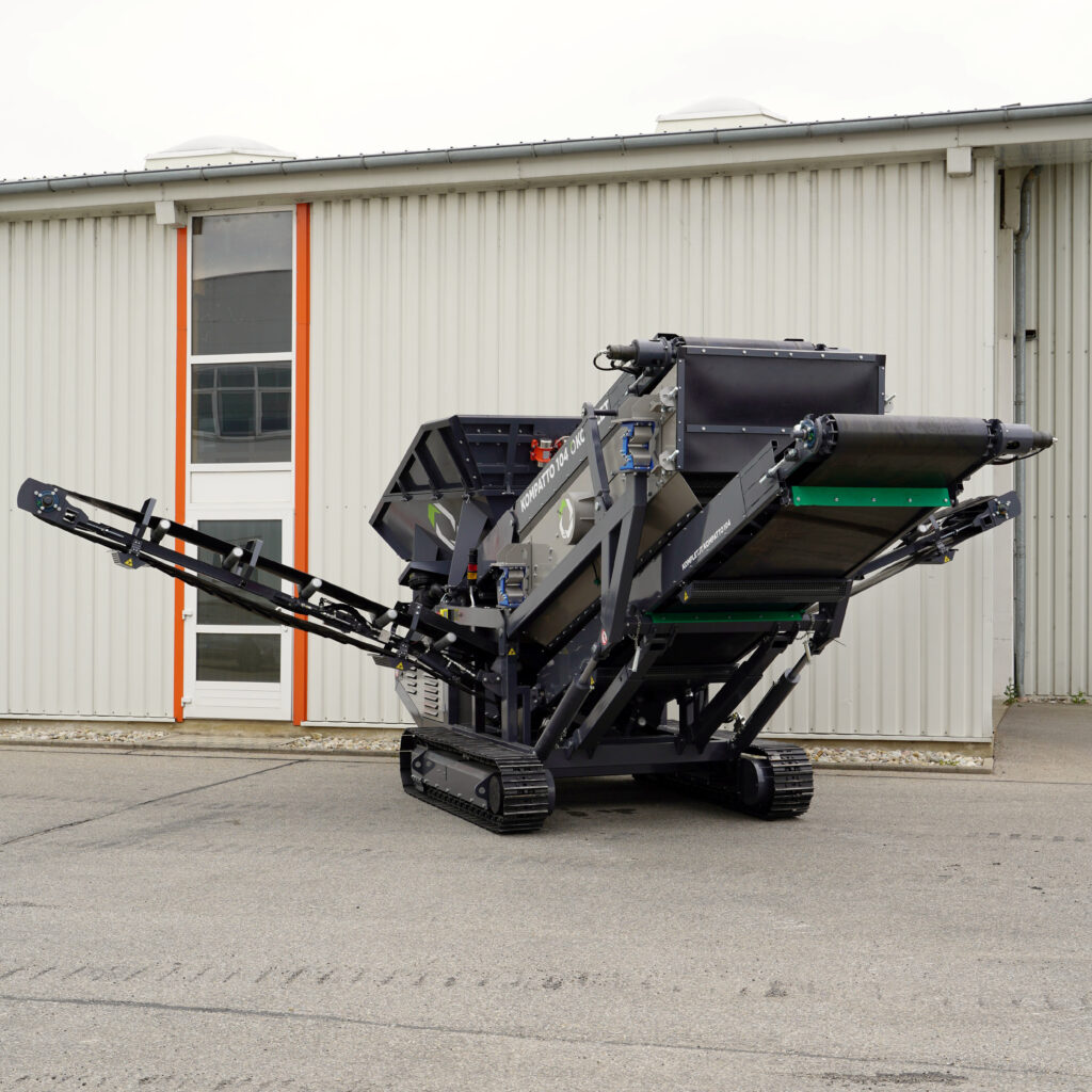 Vor einem hellen Industriebau steht ein schwarzer Kompatto 104-4 mit Raupenfahrwerk, Auslegern und Förderband für Recyclingmaterial im Freien.
