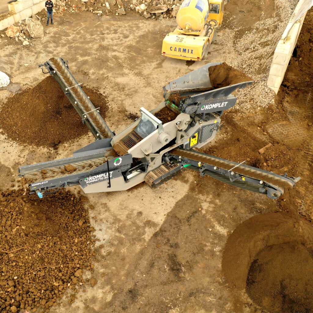 Auf einer Baustelle verteilt ein Komplet Kompatto 5030 grobes Erdmaterial, während ein gelber CARMIX-Mischer im Hintergrund zwischen Fels und Schotter steht.