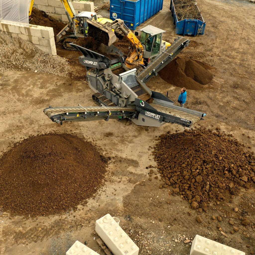 Ein gelber Bagger lädt Erde in eine graue Komplet Kompatto 5030, daneben liegen große Schüttguthaufen und blaue Container auf dem Gelände.