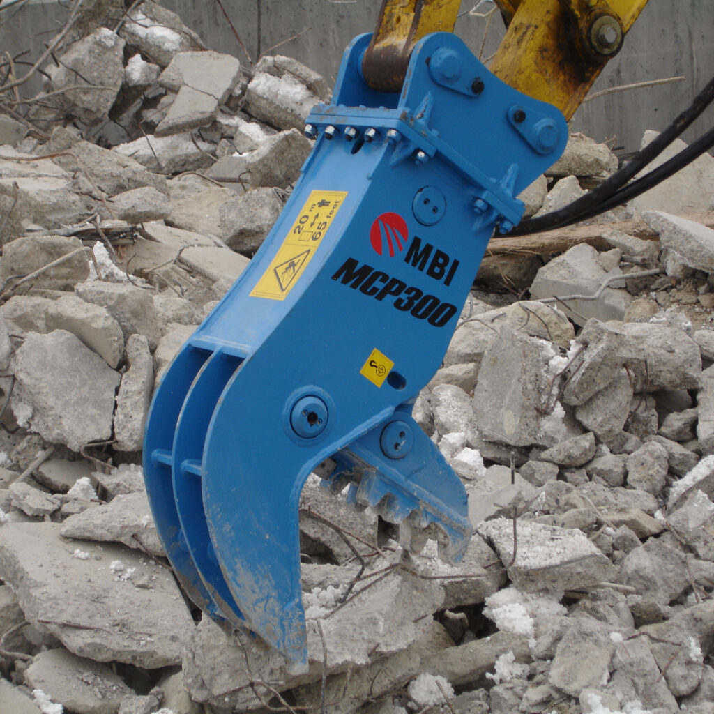Der blaue MBI MCP300 greift mit seinem Schneidwerk in einen Haufen aus Betonbruch, während gelbe Hydraulikarme und Schläuche das schwere Gerät führen.