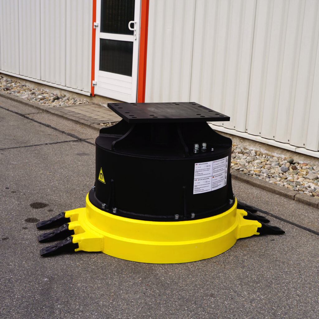 Ein schwarzes Anbaugerät mit gelbem Sockel steht vor einer weißen Industriehalle, daneben liegen Pflastersteine und ein schmaler Gehweg aus Asphalt.