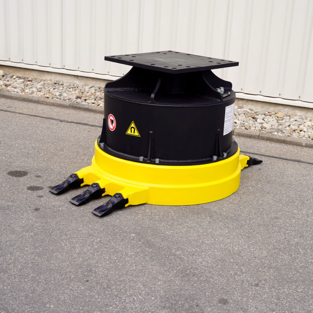Ein gelb-schwarzes Maschinenbauteil mit rechteckiger Stahlplatte, Warnsymbolen und drei Zähnen steht auf Asphalt vor einer hellen Wand im Freien.