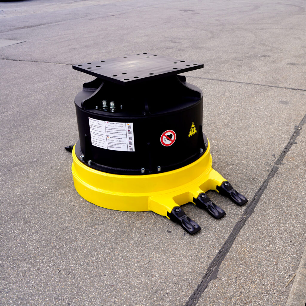 Ein schwarz-gelber Drehkranz mit Stahlplatte und drei Zinken steht auf grauem Asphalt; auf dem Gehäuse sind Warnhinweise und Anschlüsse sichtbar.