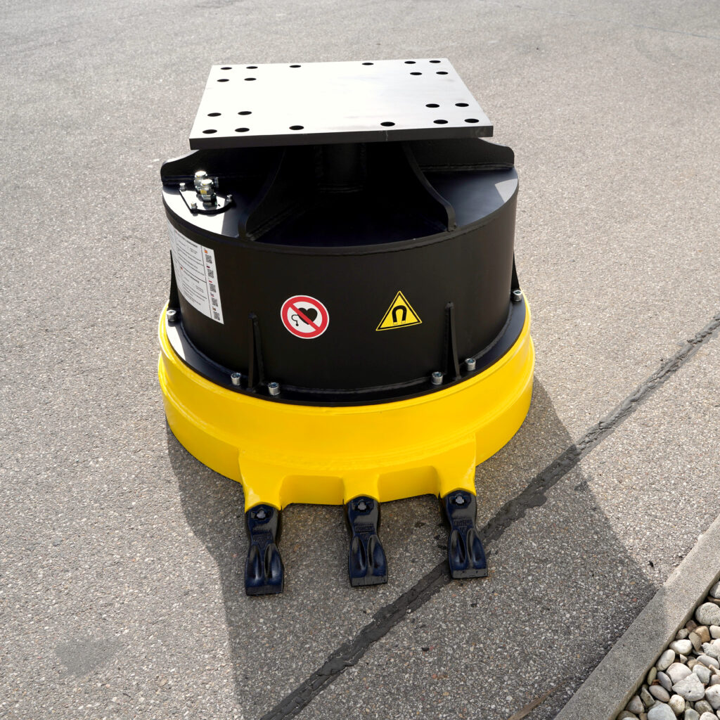 Ein gelb-schwarzes Anbaugerät mit zylindrischem Gehäuse, Metallplatte und Warnaufklebern steht auf Asphalt im Freien bei hellem Sonnenlicht.