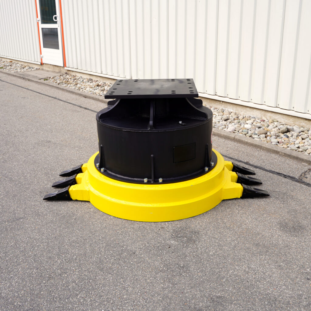 Ein gelb-schwarzes Anbaugerät steht auf Asphalt vor einer hellen Industriehalle, mit massiver Rundform, Montageplatte und seitlichen Zähnen.