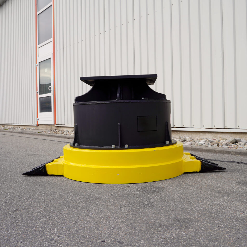 Ein gelb-schwarzes, rundes Bauteil steht vor einer hellen Industriehalle auf Asphalt; seitlich sind kleine Metallteile und ein hoher Aufsatz zu sehen.