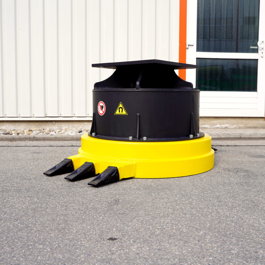 Schwarzer, zylindrischer Anbau mit gelbem Sockel und drei Zinken steht vor einer hellen Industriehalle mit orangefarbener Strebe.