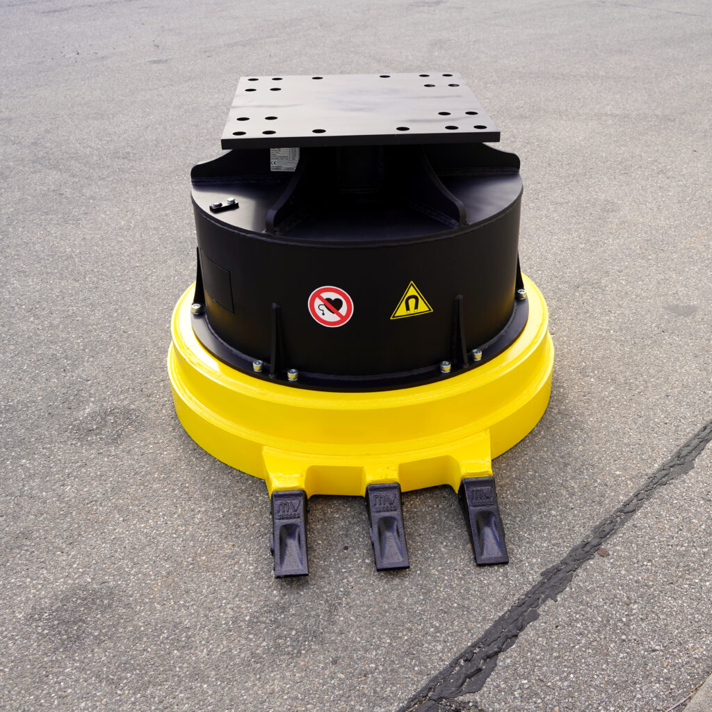 Ein gelb-schwarzes Anbaugerät mit runder Basis, Stahlplatte und drei Zinken steht auf Asphalt und zeigt Warnsymbole sowie Befestigungspunkte.
