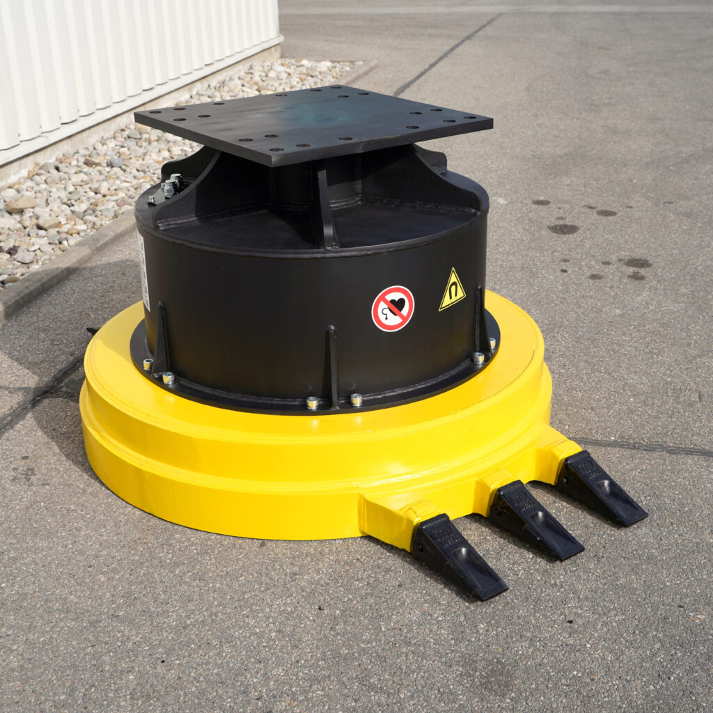 Ein gelb-schwarzer Maschinenaufsatz mit großem Stahlflansch, drei Zinken und Warnsymbolen steht auf Asphalt neben einer hellen Hallenwand.