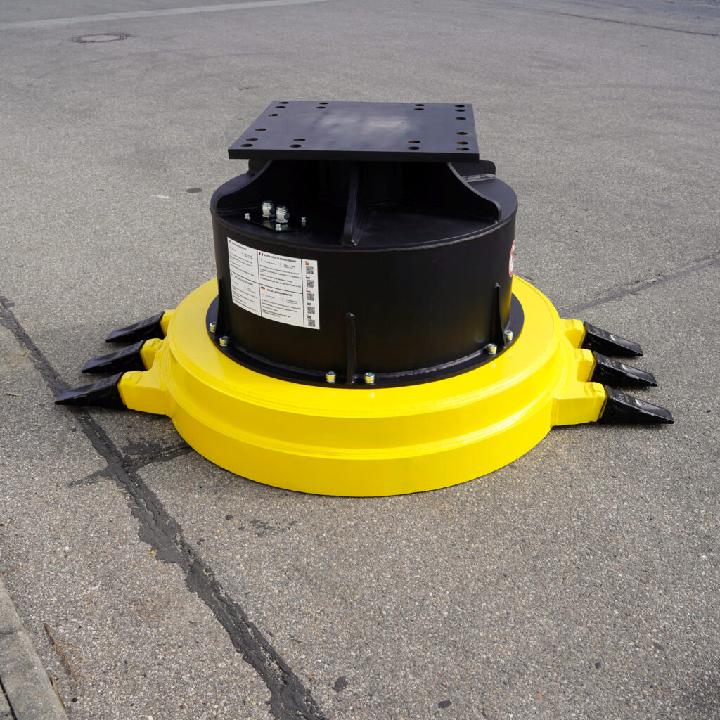 Schwarzes, zylindrisches Maschinenbauteil mit gelbem Ring und Lochplatte oben steht auf Asphalt, umgeben von Metallbolzen und Warnhinweisen.