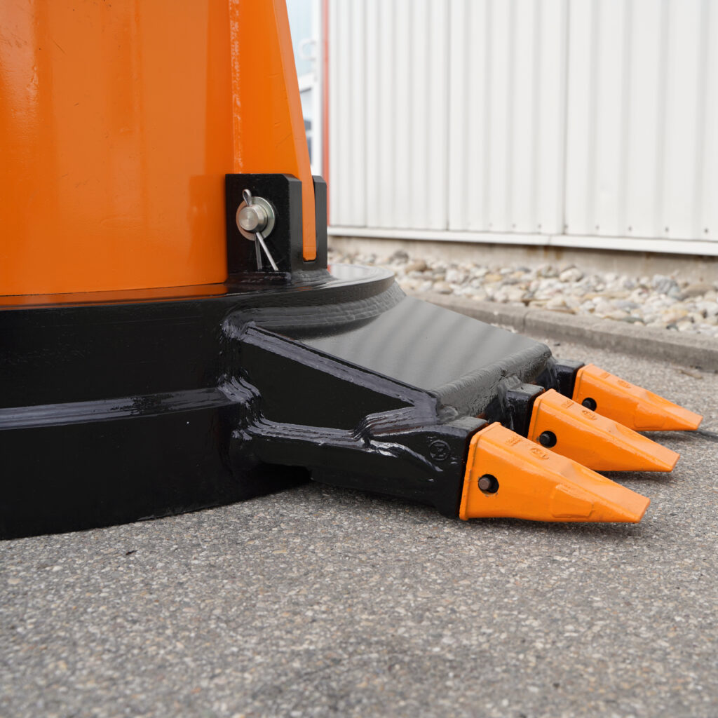 Nahaufnahme eines orange-schwarzen Baggeranbauteils mit drei orangefarbenen Zahneinsätzen und Metallbolzen vor einer hellen Wand.