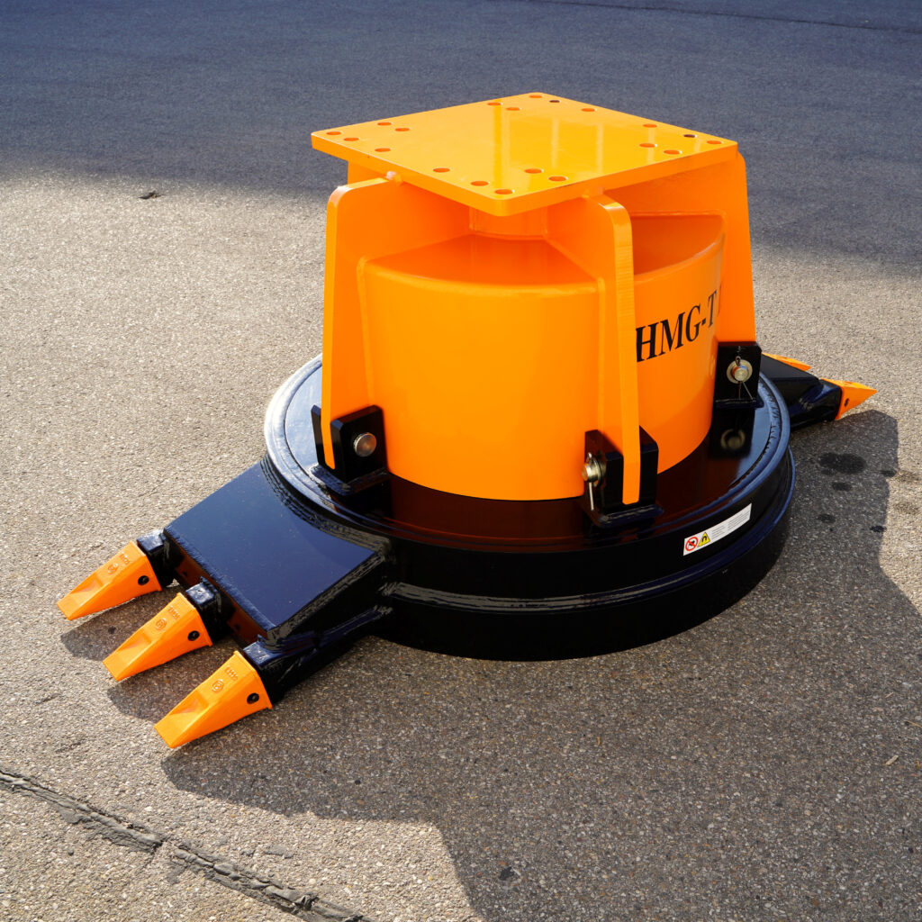 Ein orange-schwarzes Maschinenanbaugerät mit mehreren Zähnen steht auf Asphalt, daneben sind die Verschraubungen und die runde Grundplatte sichtbar.