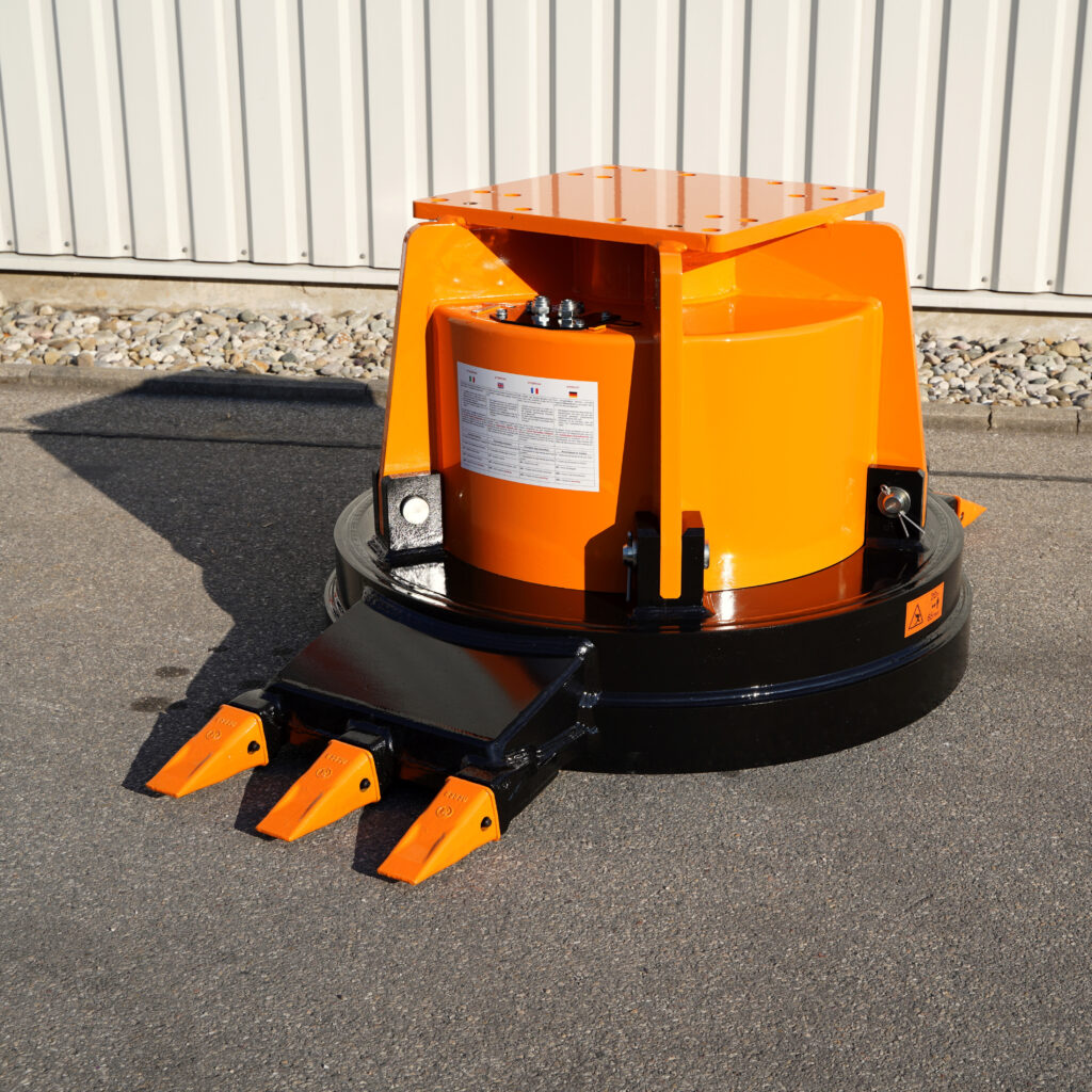 Ein orange-schwarzer Brecheraufsatz mit drei spitzen Zähnen steht vor einer hellen Blechwand auf Asphalt und zeigt robuste Stahlkonstruktion.