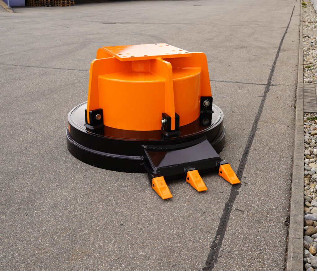 Ein orange-schwarzes Maschinenanbaugerät mit runder Grundplatte und drei Zähnen steht auf Asphalt neben einem Bordstein. Die Stahlkonstruktion wirkt massiv und robust.