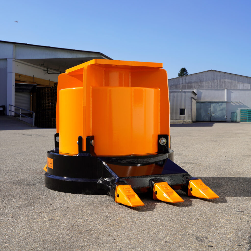 Ein orangefarbener Baggeranbau mit schwarzem Sockel und drei Zähnen steht vor Industriehallen unter klarem Himmel auf einem Asphaltplatz.