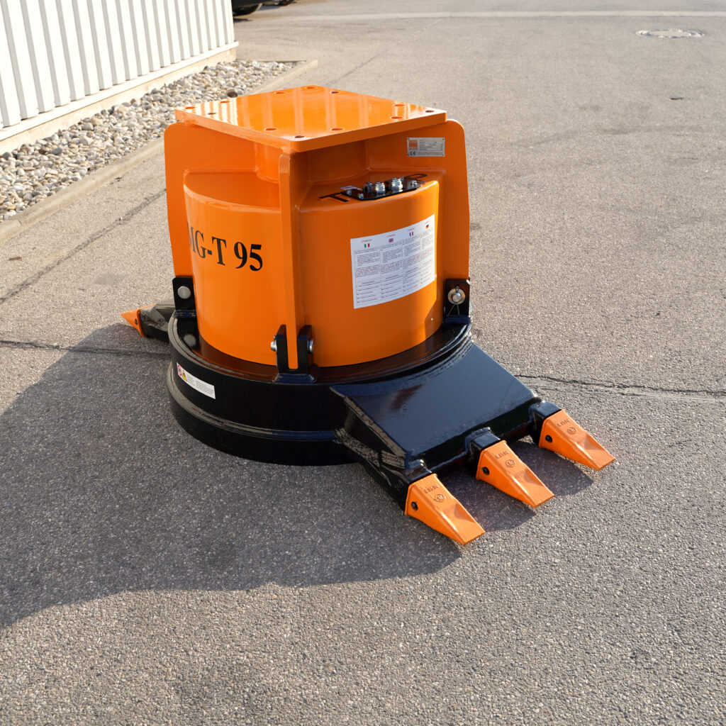 Ein orange-schwarzer Anbauverdichter mit T95-Aufdruck steht auf Asphalt vor einer hellen Hallenwand und zeigt drei scharfe Stahlzinken im Freien.