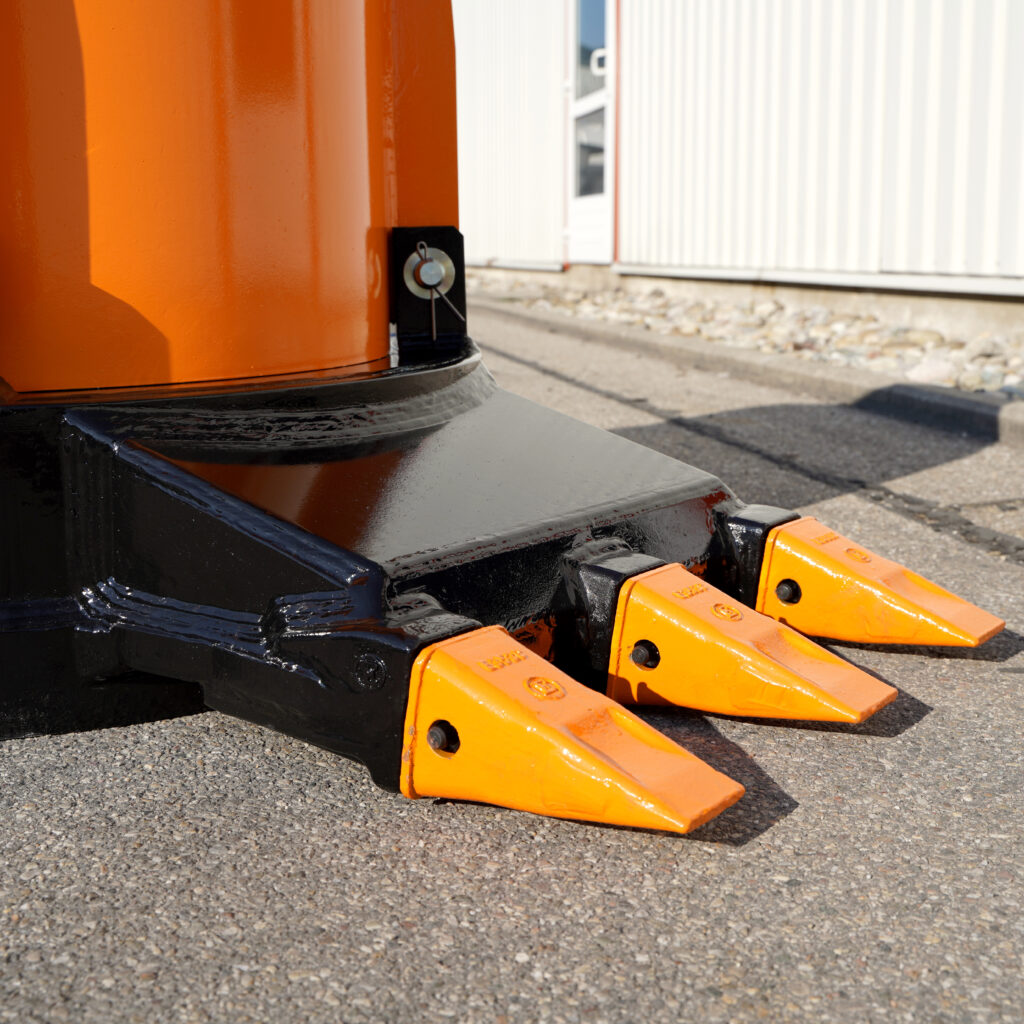 Nahaufnahme eines orange-schwarzen Baggeranbaugeräts mit drei gezackten Zähnen auf Asphalt vor einer hellen Industriehalle im Sonnenlicht.