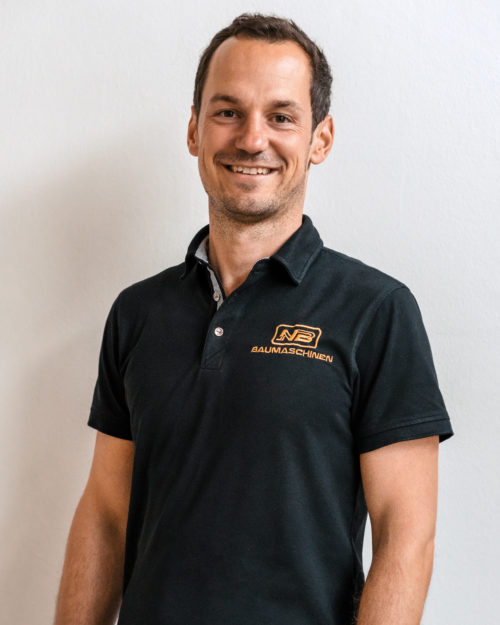Niklas Brimm steht vor einer hellen Wand und lächelt in einem schwarzen Polohemd mit orangefarbenem NB-Baumaschinen-Logo auf der Brust.