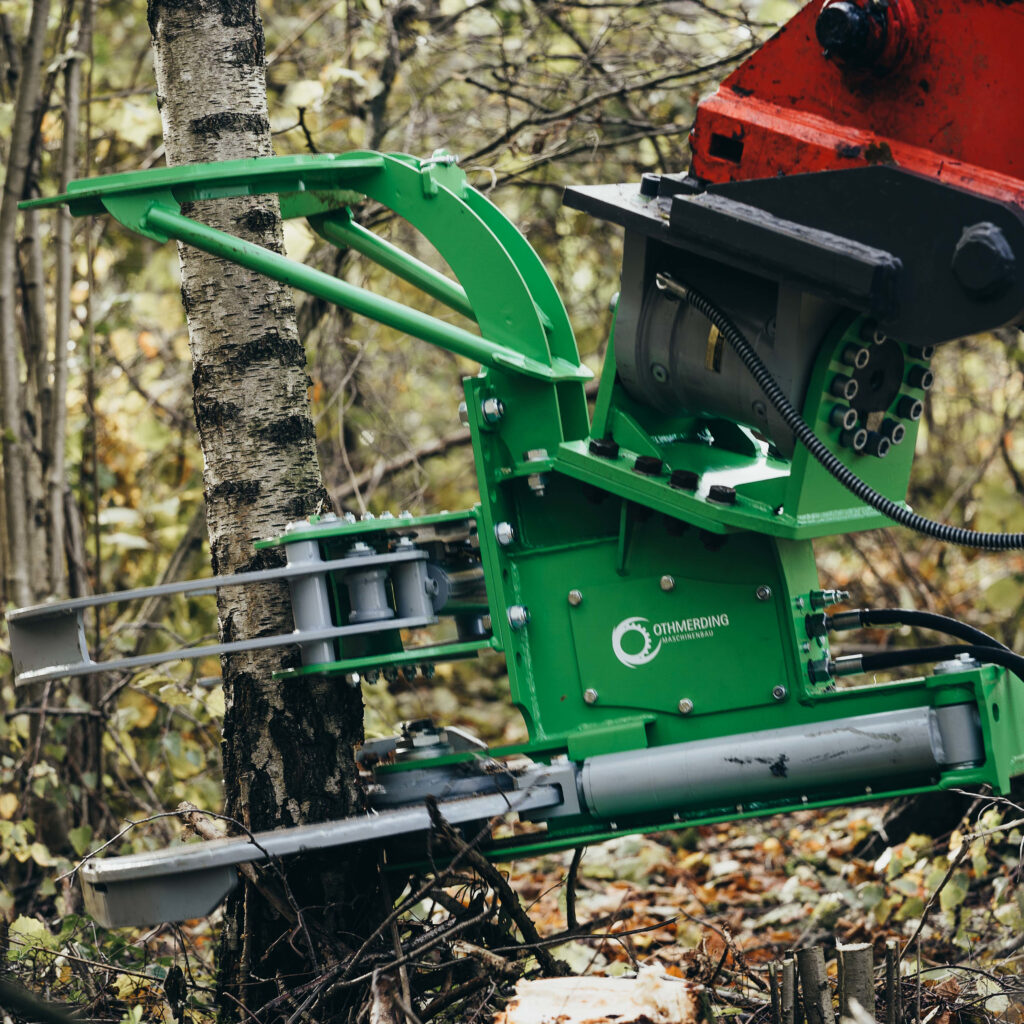 Ein grünes Othmerding-Baumscheren-Aggregat greift einen Birkenstamm im Wald, während rote und schwarze Hydraulikteile rechts im Fokus stehen.