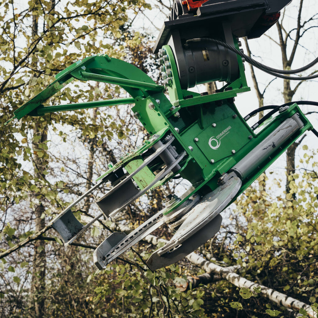Grüne Othmerding-Maschinenbau-Anbaugerät mit mehreren Greifarmen und Hydraulikzylindern vor belaubten Bäumen, schräg von unten aufgenommen.