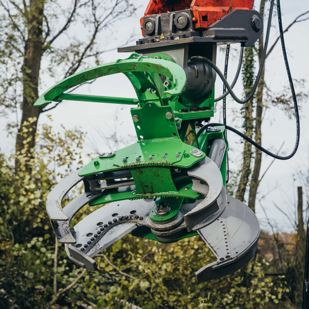 Vor unscharfen Bäumen hängt eine grüne Othmerding-Baumschere mit grauen Greifarmen, Bolzen und Hydraulikschläuchen im Freien.