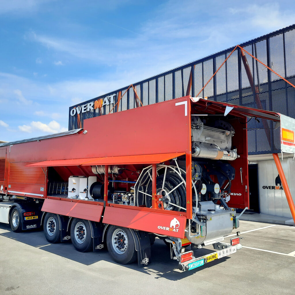 Ein roter Overmat-Maschinenaufbau steht auf einem Lkw im Freien vor blauem Himmel, mit geöffneten Klappen, Schläuchen und sichtbar verbauter Technik.