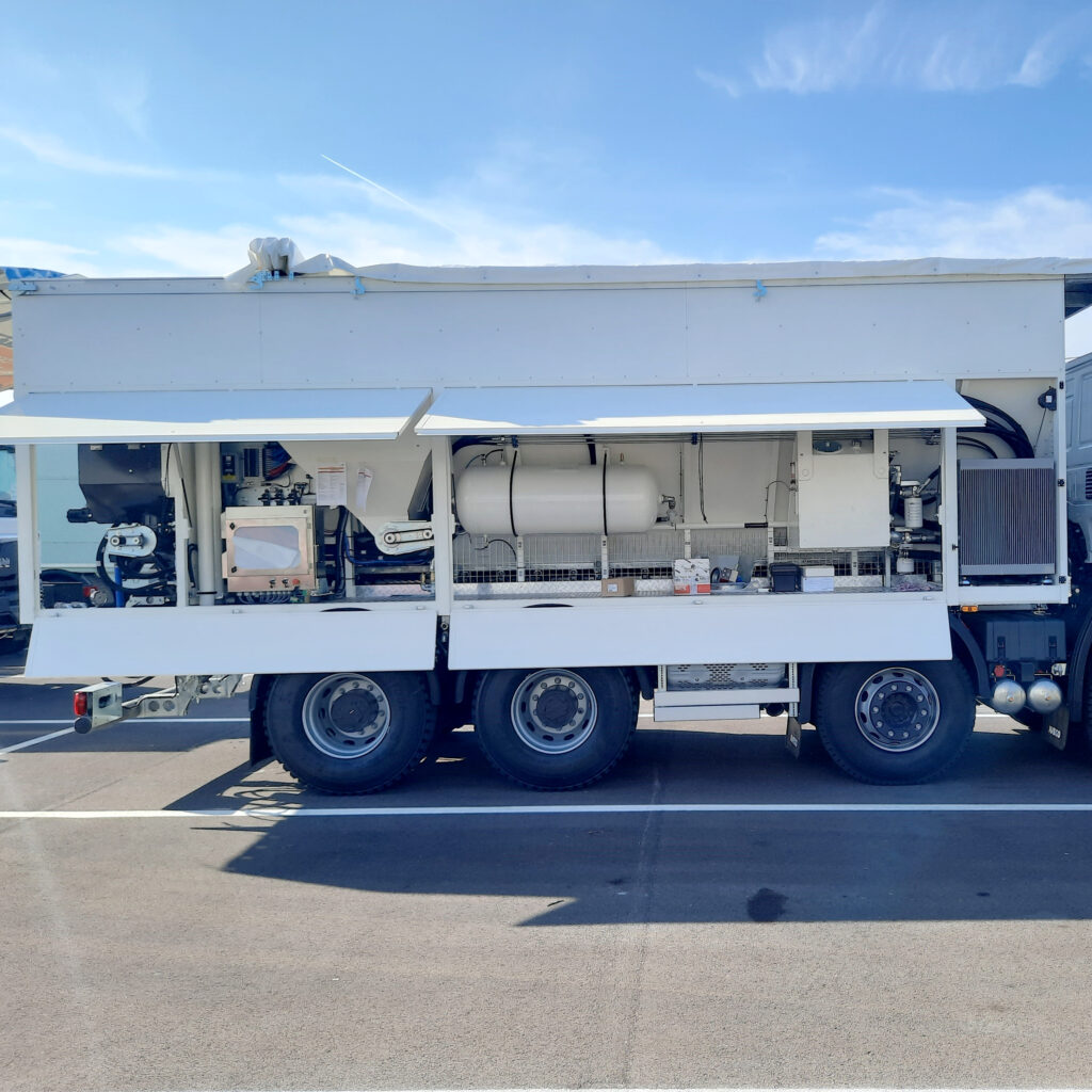 Eine große weiße Overmat-Anlage mit freiliegenden Hydraulik- und Technikkomponenten steht seitlich auf einem Lkw-Fahrgestell unter klarem Himmel.