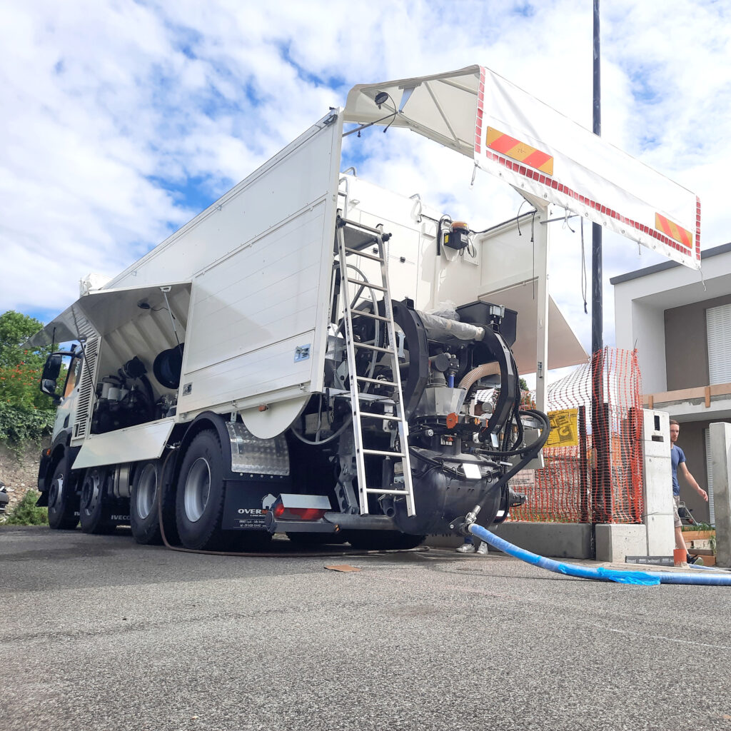 Ein weißer Overmat-Betonmischer mit ausgefahrenem Ausleger steht an einer Baustelle, daneben liegen Schlauch und Leiter unter blauem Himmel.