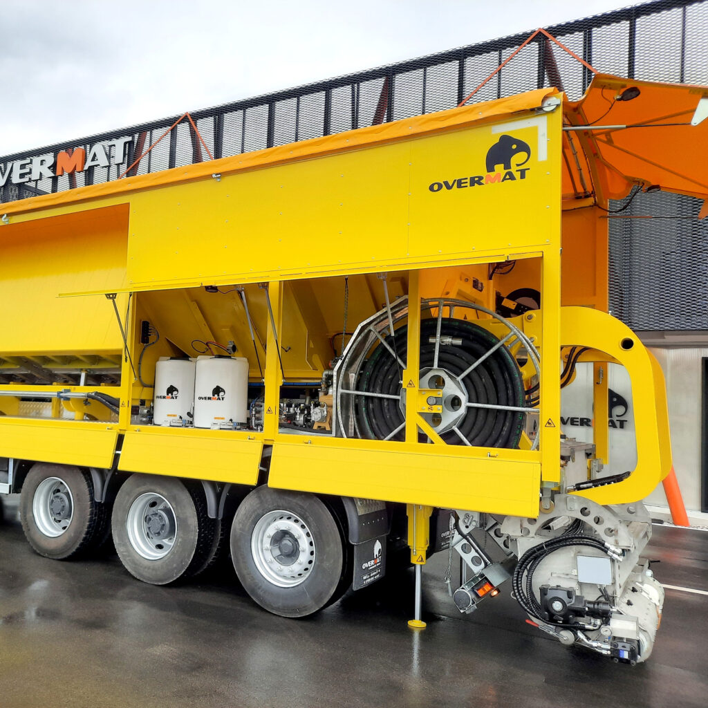 Ein gelber Overmat-Auflieger mit großen Schlauchrollen, sichtbaren Hydraulikbauteilen und orangefarbener Abdeckung steht vor einer Metallfassade.