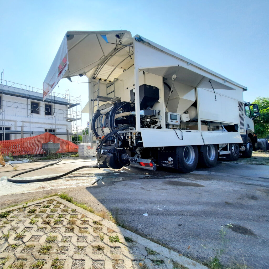 Ein weißer Overmat-Sattelzug mit aufklappbarem Aufbau, Schläuchen und Technik steht neben einem Baugerüst auf einer Baustelle unter klarem Himmel.